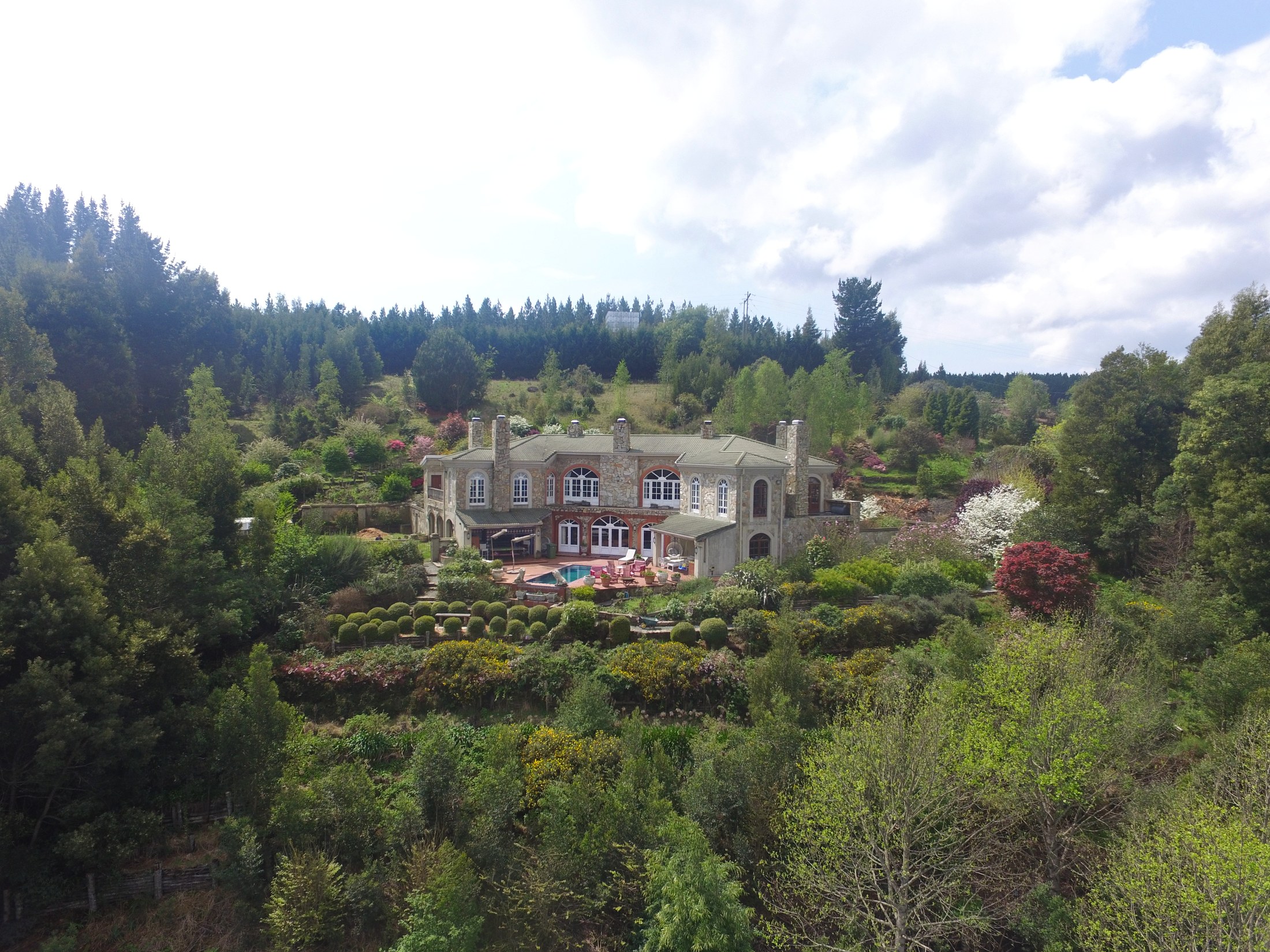 Wide view of the Evergarde stone manor, pool terrace, and gardens under clear sky